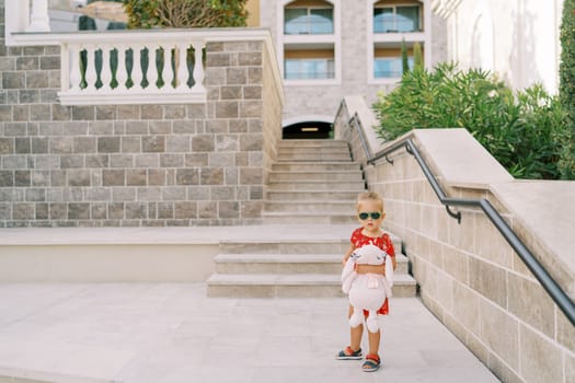 Little girl with a pink toy hare stands near the stairs of an apartment building in the garden. High quality photo