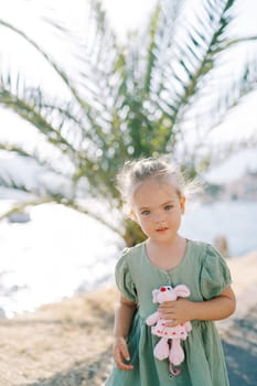 Little girl with a soft toy in her hand stands on the seashore near a green palm tree. High quality photo