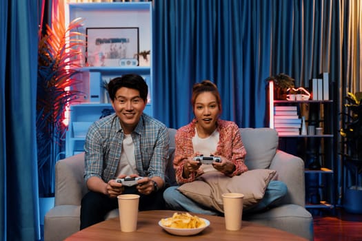 Sitting sofa of happy smiling couple playing video game control by joystick fronted snack and drinks competing challenge level with funny at modern neon blub light comfy living room at home. Infobahn.