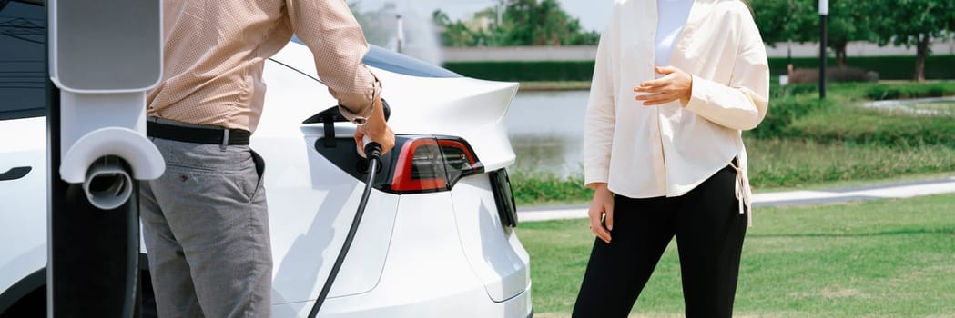Young couple recharge EV car battery at charging station connected to power grid tower electrical industrial facility as electrical industry for eco friendly vehicle utilization. Panorama Expedient