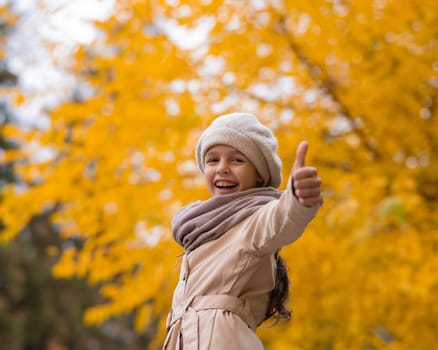 Happy caucasian girl in a beige coat and beret walks in the park in autumn. Schoolgirl showing thumbs up