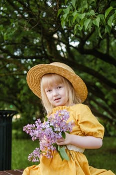 A little girl in a yellow dress and straw hat wearing a bouquet of lilacs. A walk in a spring park, blossoming lilacs