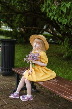 A little girl in a yellow dress and straw hat wearing a bouquet of lilacs. A walk in a spring park, blossoming lilacs