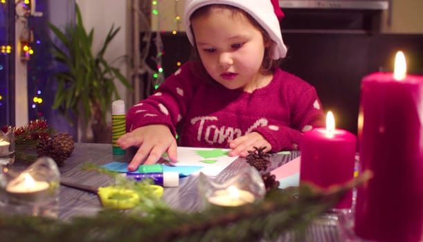 Children handicraft. Little girl glues New Year card. Table with Christmas decorations. French text on a sweater - falls