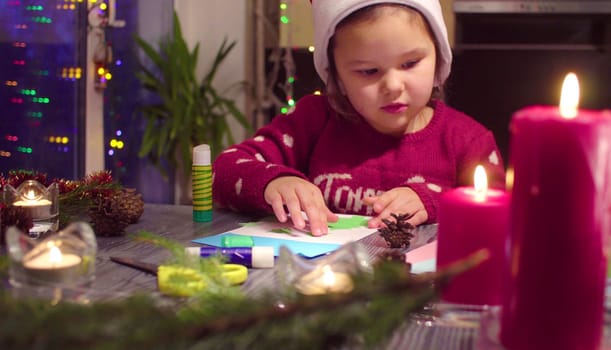 Children handicraft. Little girl glues New Year card. Table with Christmas decorations. French text on a sweater - falls