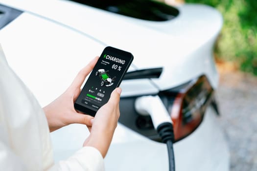 Young woman use smartphone to pay for electricity at public EV car charging station green city park. Modern environmental and sustainable urban lifestyle with EV vehicle. Expedient