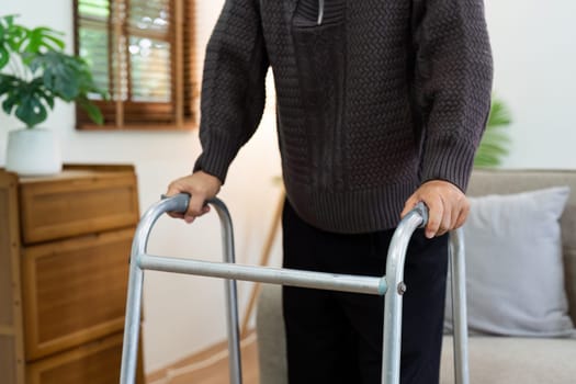 A man in a black sweater is using a walker. The walker is silver and has a handle on each side. The man is standing in a living room with a couch and a potted plant