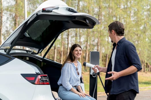 Lovely young couple recharging battery for electric car while relaxing and sitting on the trunk during road trip travel EV car in autumnal forest. Eco friendly travel on vacation during autumn. Exalt