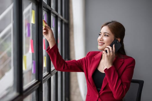 Asian businesswoman talking on the phone talking about a work meeting It's near the wall with a sticky note..