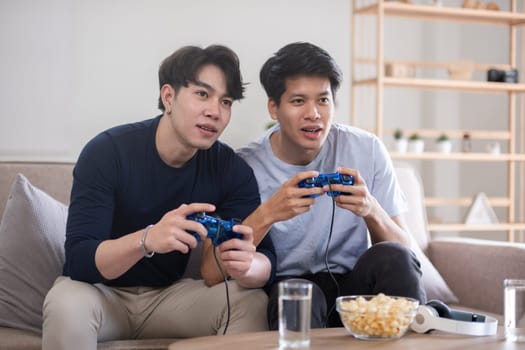 A fun lgbt male couple sits happily playing a pc game with a console joystick together..