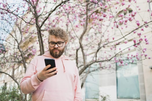 Spring day. Bearded man in pink shirt talking by phone. Spring pink sakura blossom. Handsome young man with smartphone. Fashionable man in trendy glasses. Bearded stylish man. Male fashion.