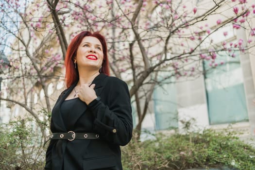 Fashion outdoor photo of beautiful woman with red curly hair in elegant suit posing in spring flowering park with magnolias tree. Copy space and empty place for advertising text.