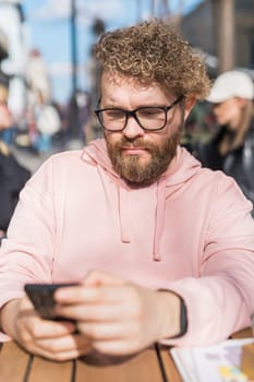 Portrait of cheerful millennial blogger with modern cellphone technology enjoying freelance lifestyle, happy hipster guy in optical eyewear using mobile phone in street cafe.