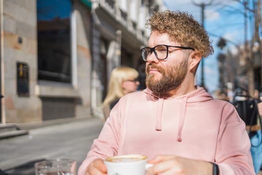 Young bearded man having breakfast at table of street cafe on spring day, drinking warm cappuccino and enjoy morning