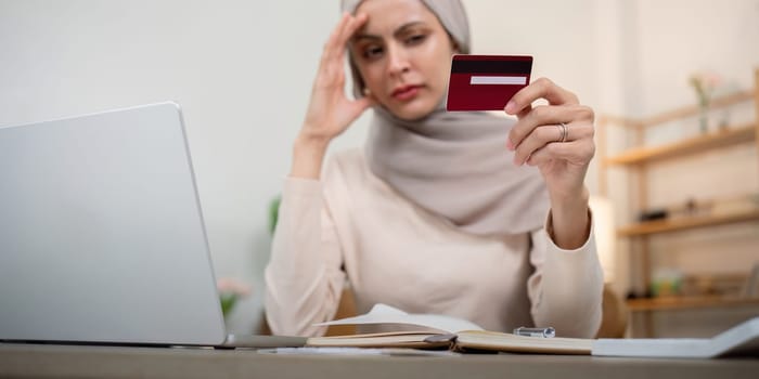 Credit card problem. Worried and stress young muslim woman in hijab sitting in living room working and make purchase in online store on laptop.