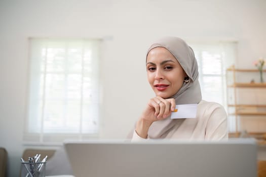 Young happy muslim woman in hijab at home using laptop shopping online with credit card while sitting on desk.