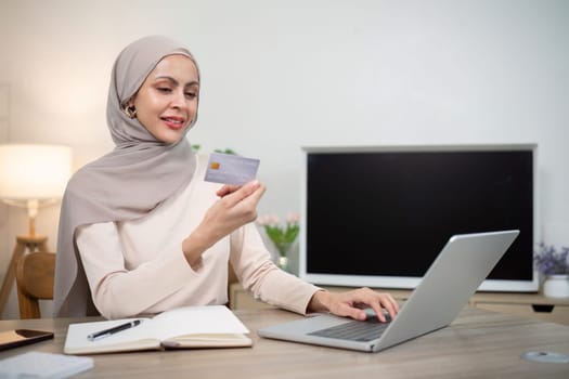 Young happy muslim woman in hijab at home using laptop shopping online with credit card while sitting on desk.