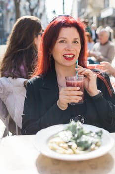 Beautiful happy woman with long red hair enjoying cocktail in a street cafe.
