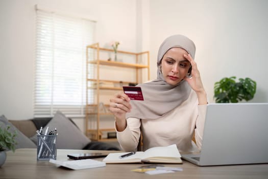 Credit card problem. Worried and stress young muslim woman in hijab sitting in living room working and make purchase in online store on laptop.