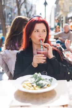 Beautiful happy woman with long red hair enjoying cocktail in a street cafe.