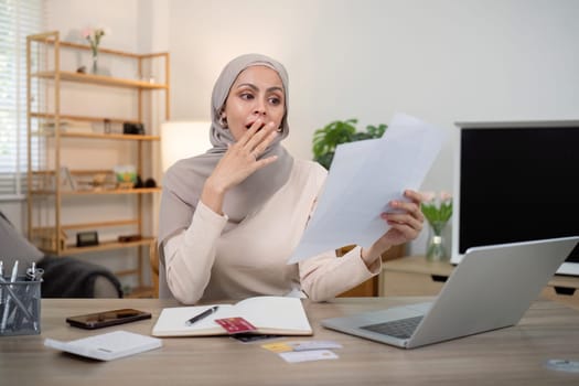 Young worried and shocked Muslim woman in hijab holding documents and calculator. Financial problems with accounts, loans, family budget at the home.