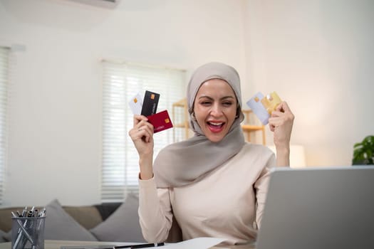 Young happy muslim woman in hijab at home using laptop shopping online with credit card while sitting on desk.