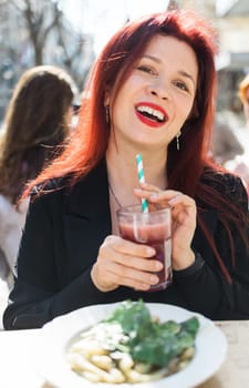 Beautiful happy woman with long red hair enjoying cocktail in a street cafe.