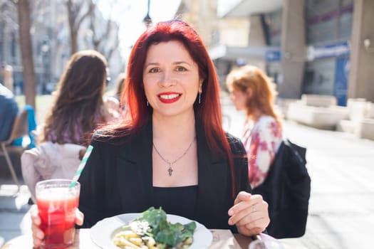 Millennial woman eating italian pasta at restaurant on the street in spring. Concept of Italian gastronomy and travel. Stylish woman with red hair.