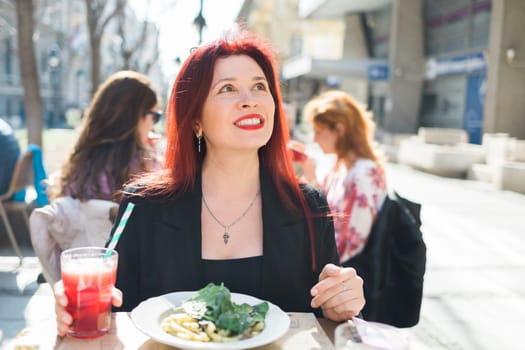 Millennial woman eating italian pasta at restaurant on the street in spring. Concept of Italian gastronomy and travel. Stylish woman with red hair.