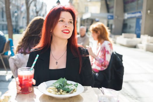 Beautiful happy woman with long red hair enjoying pasta in a street cafe