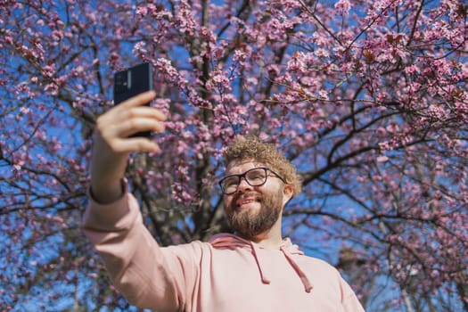 Spring day. Bearded man in pink shirt talking by phone. Spring pink sakura blossom. Handsome young man with smartphone. Fashionable man in trendy glasses. Bearded stylish man. Male fashion.