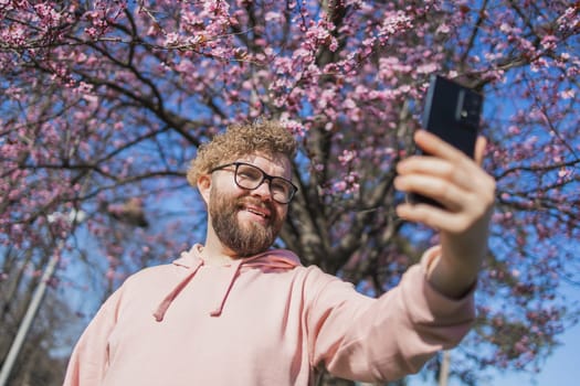 Happy curly man takes selfie against backdrop of flowering tree in spring for his internet communications. Weekend and social networks