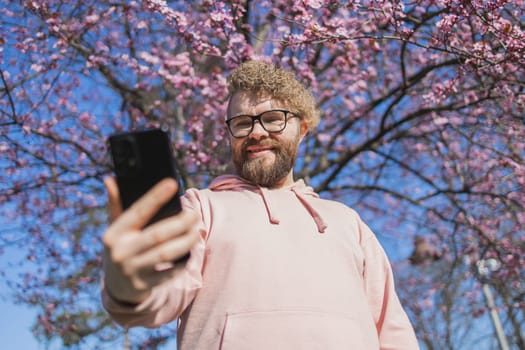 Happy curly man takes selfie against backdrop of flowering tree in spring for his internet communications. Weekend and social networks