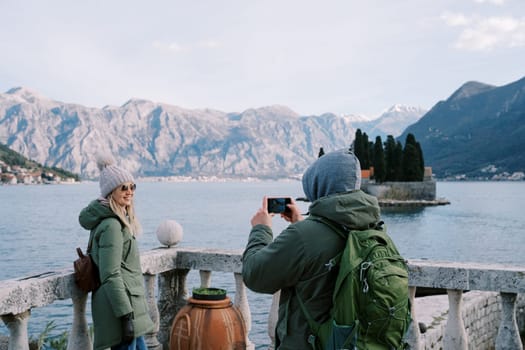 Guy takes a picture of girl with a smartphone near the balustrade on the shore of the Kotor Bay overlooking the island of St. George. Montenegro. Back view. High quality photo