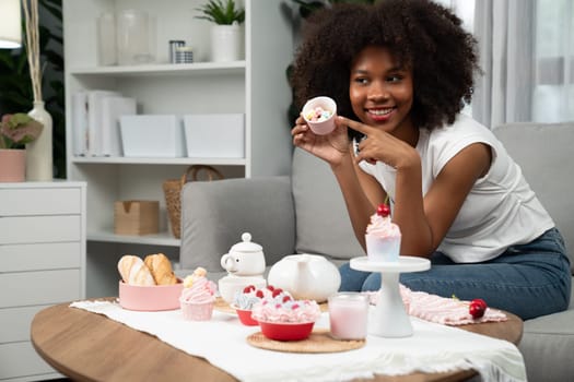 Beautiful young African blogger presenting piece of cupcake in concept showing special cuisine in pastel color. Content creating of social media with favorite sweets bakery dish. Tastemaker.