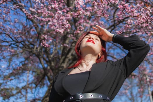 Fashion outdoor photo of beautiful woman with red curly hair in elegant suit posing in spring flowering park with blooming cherry tree. Copy space and empty place for advertising text.