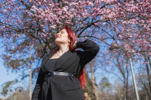 Fashion outdoor photo of beautiful woman with red curly hair in elegant suit posing in spring flowering park with blooming cherry tree. Copy space and empty place for advertising text.