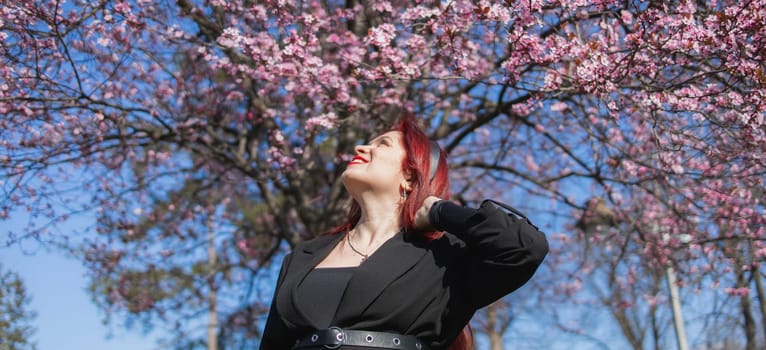 Fashion outdoor photo of beautiful woman with red curly hair in elegant suit posing in spring flowering park with blooming cherry tree. Copy space and empty place for advertising text.