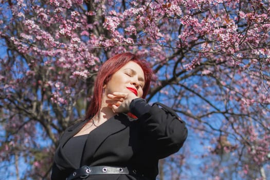 Fashion outdoor photo of beautiful woman with red curly hair in elegant suit posing in spring flowering park with blooming cherry tree. Copy space and empty place for advertising text.