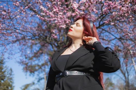 Fashion outdoor photo of beautiful woman with red curly hair in elegant suit posing in spring flowering park with blooming cherry tree. Copy space and empty place for advertising text.