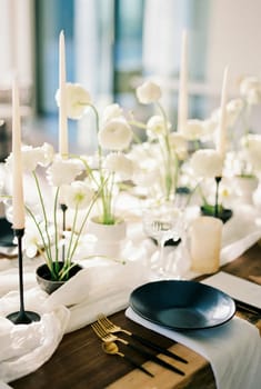 Black plates stand on a wooden table next to white bouquets of flowers on a narrow tablecloth. High quality photo