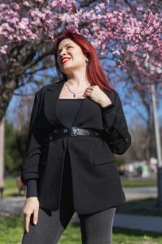 Fashion outdoor photo of beautiful woman with red curly hair in elegant suit posing in spring flowering park with blooming cherry tree. Copy space and empty place for advertising text.