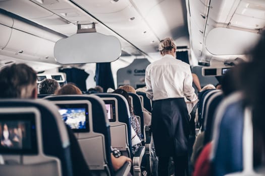 Interior of airplane with passengers on seats and stewardess in uniform walking the aisle, serving people. Commercial economy flight service concept