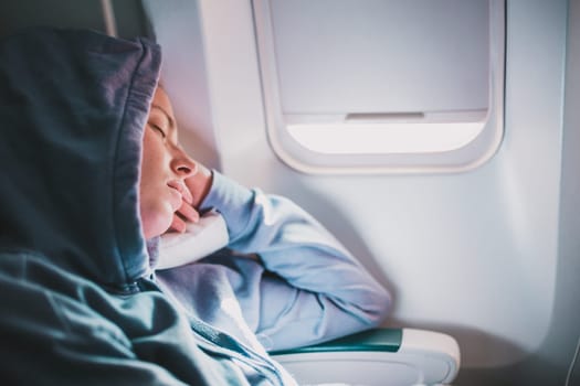 Tired blonde casual caucasian woman wearing sporty hoodie napping on seat while traveling by airplane. Commercial transportation by planes. Authentic image of real people
