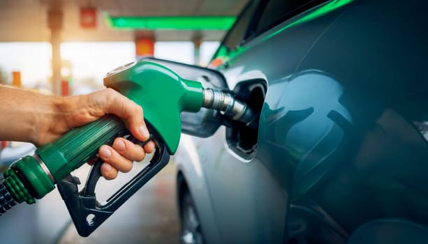 A hand grips a green fuel nozzle while refueling a car at a gas station. The nozzle is inserted into the vehicle's tank, showing the refueling process of filling up with gasoline or diesel.