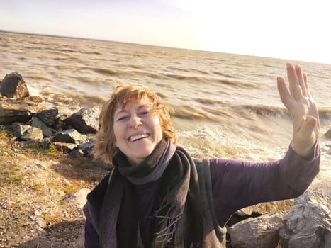 Happy cheerful middle aged woman in scarf taking selfie on nature outdoors and sun with water and waves of sea on the background