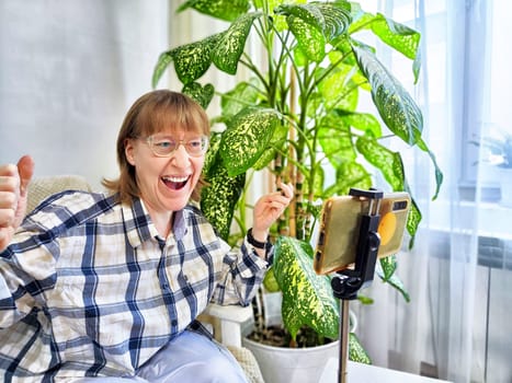 Smiling woman in glasses using a smartphone on a tripod for a video chat. A middle-aged woman posing and taking a selfie