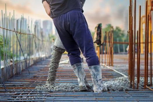 A construction site worker in rubber boots manipulates a concrete pump hose during the process of pouring concrete into a reinforcement cage.