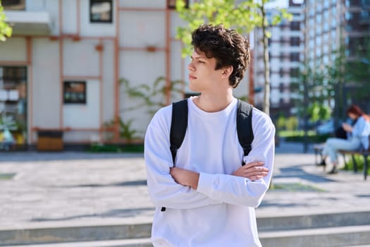 Portrait of confident smiling college student guy, young male with crossed arms with backpack looking in profile outdoor near educational building. Education, training, 19,20 years age youth concept