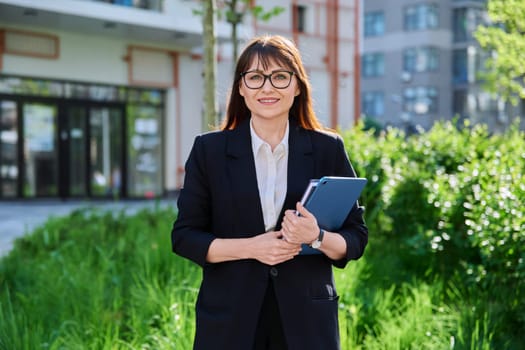 Middle-aged business woman in suit with digital tablet, outdoor building. Female teacher posing near school educational center, office worker entrepreneur accountant economist banker financier lawyer